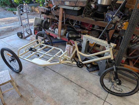 A very large and long cargo trike, freshly painted in a light cream color. It sits in a cluttered, unfinished shop space.