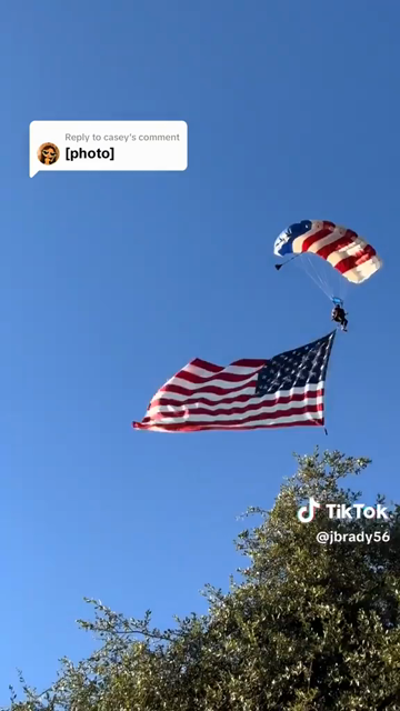 A video of a skydiver coming in to land at a public festival.  The sky is clear blue and there are a number of large trees around.  The skydiver is clearly celebrating July 4 as his canopy is US Flag colored.  He is flying a giant US Flag below him that is weighted down to keep the flag flying properly.   As he comes in the weight gets caught in some trees and he is fulcrum-ed down face first into a popup tent a vender is using.