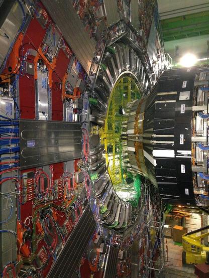 A picture of the CMS detector for the Large Hadron Collider at CERN.

A large, vertical-standing, disk-shaped conglomeration of colorful metal shapes and wiring with a large tapered cylinder covered in colorful metal shapes and wiring, adjoining perpendicularly with the disk. The whole apparatus stands several stories tall and is housed indoors with a roof distantly visible overhead.