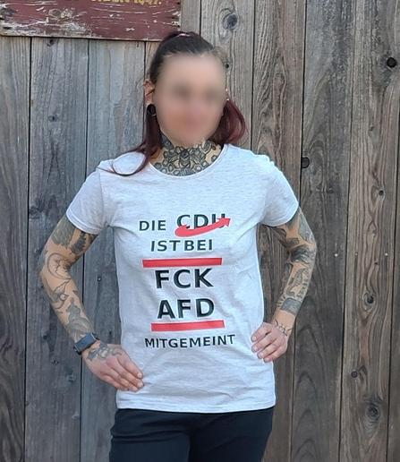 Person steht vor einer Holzwand, die Hände in die Hüften gestemmt. Sie trägt schwarze Jeans und ein graues Shirt mit dem Aufruck in schwarz und rot "Die CDU ist bei FCK AFD mitgemeint"