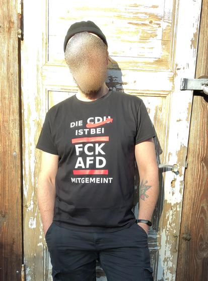 Person steht vor einer schrabbeligen Holztür, die Hände in den Hosentaschen. Sie trägt eine schwarze Hose und ein schwarzes Shirt mit dem Aufruck in weiß und rot "Die CDU ist bei FCK AFD mitgemeint"