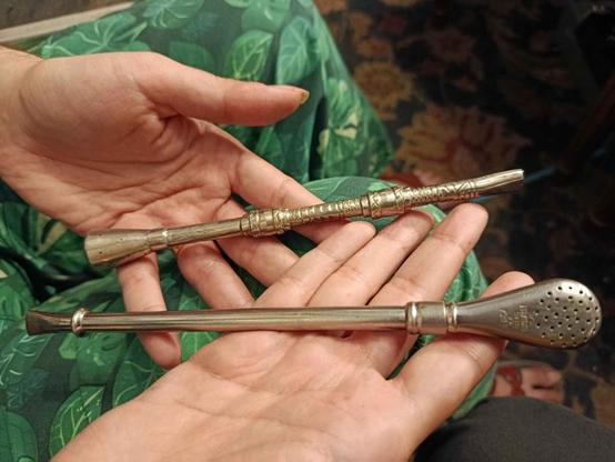 My hands holding two bombillas -- the specialized straws used for drinking yerba mate. They're two different designs, but both are a lovely sterling silver with one end looking like a regular drinking straw and one end flattened into a solid tamping end full of holes.