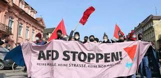 Eine Gruppe von Menschen steht auf einer Straße und hält ein großes, rosafarbenes Banner mit der Aufschrift 'AFD STOPPEN! KEINE HALLE. KEINE STRASSE. KEINE NORMALITÄT.' Mehrere Personen tragen Masken und halten rote Fahnen. Im Hintergrund sind Gebäude mit klassischer Fassadengestaltung zu sehen.