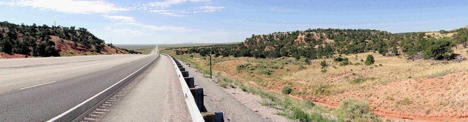 Panoramaaufnahme einer geraden, leicht abfallenden zweispurigen Landstraße, die sich durch eine trockene, hügelige Landschaft im New Mexico USA zieht. Links und rechts der Straße erheben sich mit Wacholderbüschen bewachsene Hügel mit rötlich-braunem Erdboden, dazwischen Sträucher und trockenes Gras. Am rechten Straßenrand verläuft eine Leitplanke. Die Straße führt schnurgerade bis zum Horizont, wo sich weites, flaches Grasland unter einem hellblauen Himmel mit dünnen Wolkenschleiern erstreckt.