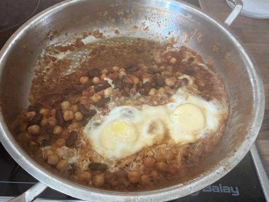 Eine Pfanne enthält eine Mischung aus Bohnen, Kichererbsen und zwei zerbrochenen Eiern, die zusammen gekocht werden. Das Gericht hat eine rötliche Sauce und etwas Bräunung an den Rändern.