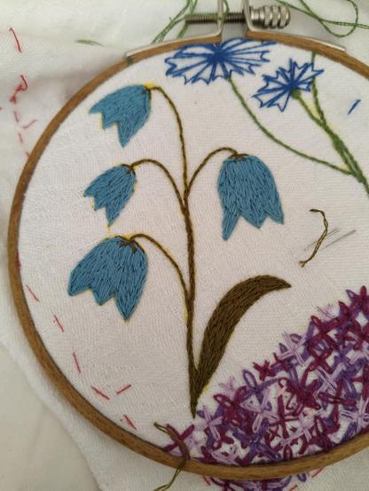 an embroidery of a bluebell plant, with 3 flowers on the left side and 1 flower and a leaf on the right side. the flowers are a light blue and the stem is a brownish green. close to it on the fabric are also embroidered some cornflowers and a lilac cluster.