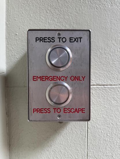 A metallic rectangular press-button panel is mounted on a textured off-white wall. Two circular metallic buttons are vertically aligned on the faceplate, with the top button labeled Press to Exit and the bottom button labeled Emergency Only and Press to Escape in red text.