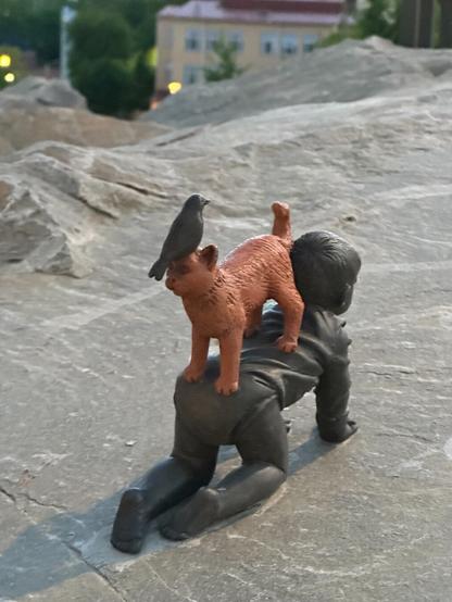 a sculpture, seemingly made of coloured metal, atop a flattened rock, in the shape of a crawling small child (in dark metal), on their back facing the other way, a standing cat (in orange), and on their head, a corvid of some sort (again in dark metal)