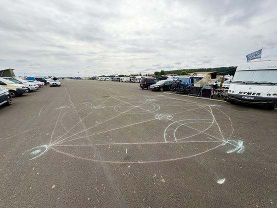 A large asphalt area features a several meter spanning chalk drawing of geometric, occult looking symbols. Numerous vehicles are parked along the sides, and gray clouds are visible in the sky.