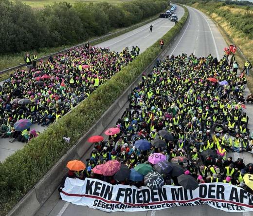 Eine große Menschenmenge, die eine mehrspurige Autobahn blockiert. Die Menschen sitzen dicht gedrängt auf beiden Fahrbahnen. 
Die vorderste Reihe hält einen Banner mit der Aufschrift "Kein Frieden mit der AfD".  Man sieht die Menschen von oben, die Aufnahme wurde von einer Brücke gemacht.