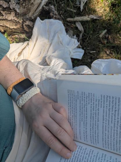 Selfie assis, centré sur mes cuisses où repose un livre ouvert. Ma main gauche est posée dessus ; au poignet, j'ai deux bracelets et une montre connectée. On aperçoit le bas de mon haut vert sauge et ma jupe plissée écrue sur un sol jonché de copeaux de bois