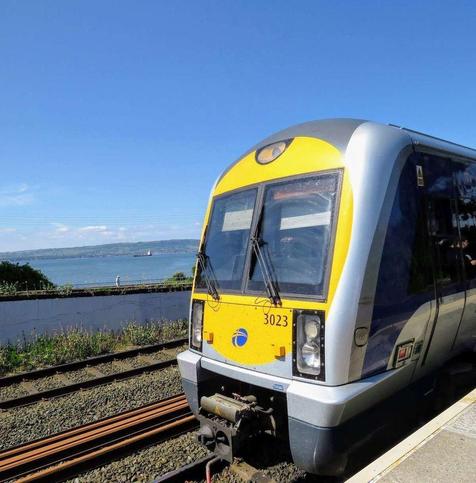 A close-up, angled shot of a silver and blue commuter train with a bright yellow front, numbered 3023, stopped at an outdoor platform. The train tracks run parallel to a low white wall, with a vast blue bay and distant green hills visible under a clear, bright sky.
