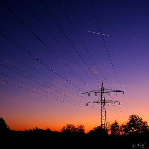 A scene at dawn. The sky transitions from dark blue at the top to blue and then to orange towards the bottom. A high-voltage power line runs across the picture from the top left to the bottom right. A single high-voltage pylon stands to the right of centre. The silhouettes of bushes and trees can be seen at the bottom edge.