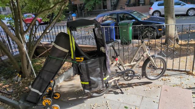 A cargo bike with a dolly attached to the back, the dolly is tied on with some old inner tube and is being used to carry a large pavilion style tent.