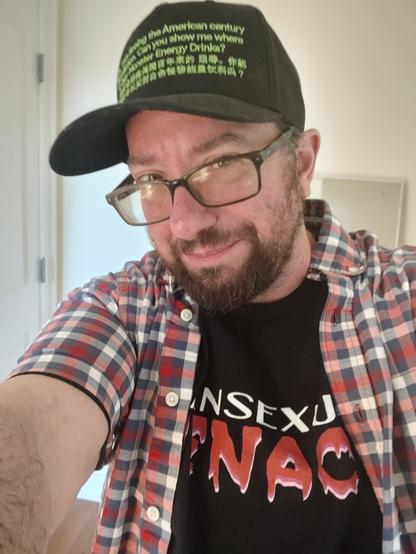 Selfie of Evan Urquhart, a bearded man with dark hair and black rimmed glasses, in a black transsexual menace t shirt, an open red white and blue short sleeve button up, and a black hat with green lettering saying "hello, I am fleeing the American century of humiliation. Can you tell me where to buy Monster energy drinks?"
