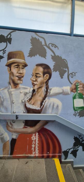 A mural painted on a light blue wall depicts two people surrounded by grey vine and leaf patterns. One person wears a tan hat and a white buttoned jacket while looking toward the second individual. The second person has long, dark braids, a white top with lace at the neckline, and a red skirt featuring vertical white decorative elements. This person holds an upright green bottle in an outstretched hand on the right side of the image. A grey railing containing small black text "TFS" cuts across the lower portion of the mural above concrete stairs.