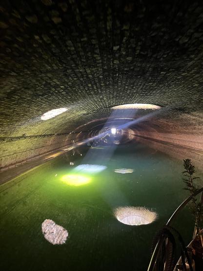 Une photo dans le tunnel du canal st Martin, à peu près au même endroit que dans le pouet précédent, mais dans des conditions de lumière différente. On voit:
- au fond, le bout du tunnel au port de l’arsenal, et la lumière fait un trait oblique sur l’objectif.
- la lumière directe du soleil par l’ouverture circulaire du plafond de la voûte, qui éclaire aussi la poussière en suspension et fait un rayon bleuté jusqu’à l’eau.
- le reflet dans l’eau de l’ouverture circulaire
- le plafond illuminé par le reflet à la surface de la lumière directe
- le reflet dans l’eau de ce deuxième cercle lumineux
- le fond de l’eau sous la lumière directe, qui fait une tache verte brillante
