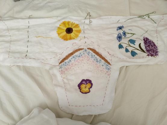 an unsewn white linen coif with embroidered flowers. so far it has a sunflower, cornflowers, bluebells, a lilac cluster, a purple and yellow viola, two reeds of grass, and chevrons full of blue, pink and white forget-me-nots.