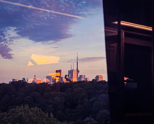 The CN Tower and downtown buildings at sunset. Trees in the foreground. The right side of the photo is dark from the structure of the viaduct. The interior of the subway car is reflected in the glass.