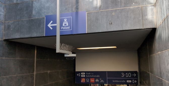 Treppenabgang, mittig eine Kamera, die Treppe ist nicht zu sehen. Im Hintergrund, unten, ein Schild, das auf die Gleise 3-10 und Wege in die Stadt hinweist
