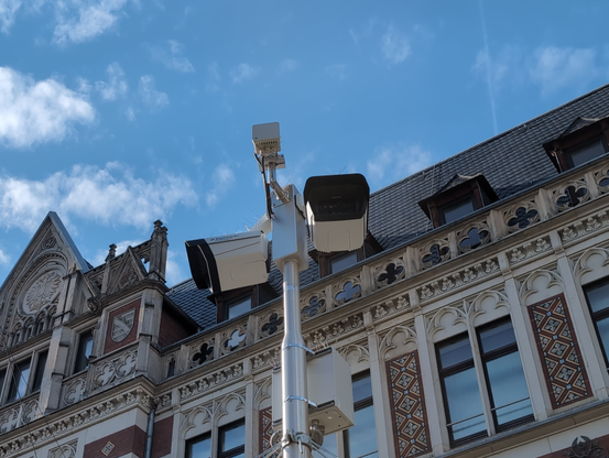 Mast mit zwei mächtigen Kameragehäusen und kleinen Schränken mit Technik. Dahiner ein historisches Gebäude