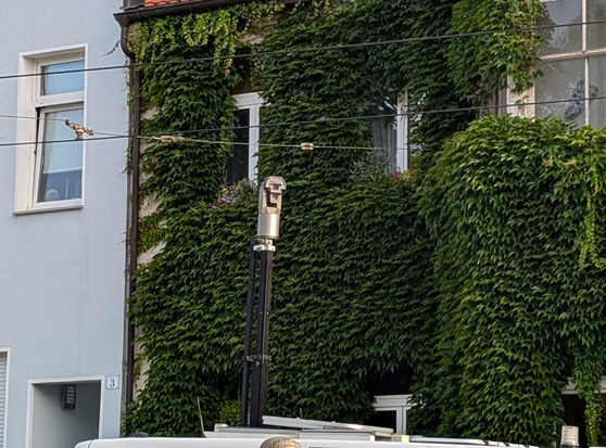 Ein ähnlicher Transporter mit senkrecht nach oben ausgefahrener Kamera, im Hintergrund ein Wohnhaus mit bewachsener Fassade