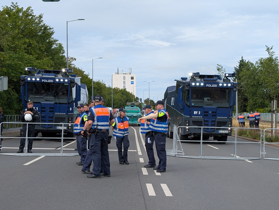 Links und rechts je ein blau-grauer Wasserwerfer. Im Hintergrund ein grüner Polizeipanzer. Davor Hamburger Gitter und eine Gruppe Polizist*innen
