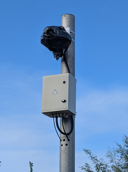 Mast an dem oben etwas mit schwarzer Plastikfolie verhüllt wurde, dadrunter ein kleiner Schrank mit Technik. Dahiner überwiegend blauer Himmel