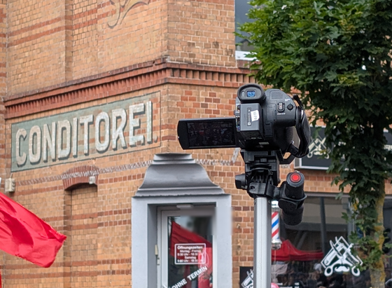 Mittig ein Camcorder am Stock, dahinter ein Gebäude an dem seitlich Conditorei steht