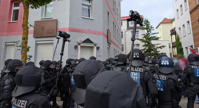 eine größere Gruppe Polizist*innen, alle behelmt und dunkel uniformiert.  Drei halten Camcorder am Stock hoch
