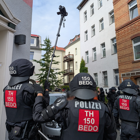 Drei Polizisten mit roten Quadrat auf ihrer Uniform, Aufschrift TH 150 BEDO. Einer mit Camcorder am Stock