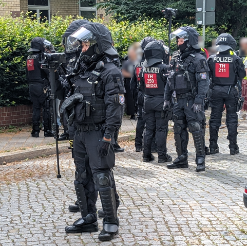 Ein behelmter Polizist hält einen Camcorder am Stock auf Hals-Höhe vor sich, der Stock hängt in der Luft