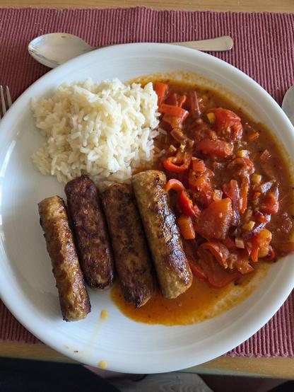 Weißer Teller mit Reis, Paprikagemüse und Cevapcici. Darunter ein rotes Tischset, Besteck um den Teller drapiert.