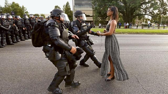 Black woman in a dress standing tall and poised facing the left while multiple cops in riot gear run forward to grab her by the arm to arrest her during Black Lives Matter protests