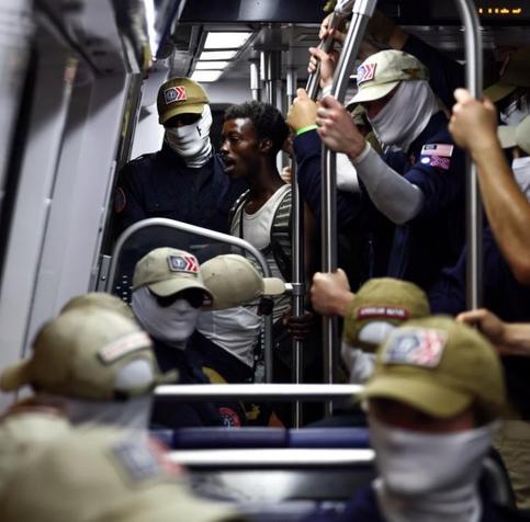 Washington DC subway car full of Patriot Front fascists with face coverings and matching surrounding a single black man facing towards the door. A patch with the Confederate flag can be seen on the left shoulder of one of them.