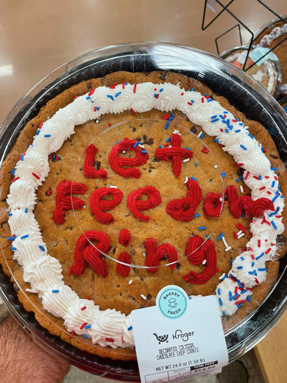 A giant cookie from Kroger inside a covered plastic tray. There’s a white frosting ring around the outside perimeter of the cookie, and inexpertly formed red frosting letters in the center which spell “Let Feedom (sic) Ring.” Red, white, and blue sprinkles are scattered around the cookie and icing, but most are strangely concentrated on the lower right - the 4:00-5:00 zone of the outer perimeter.
