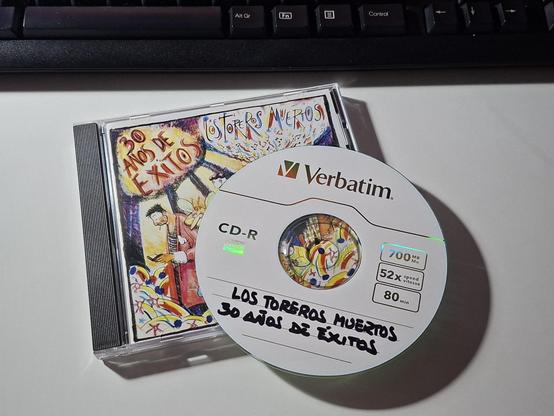 La imagen muestra un CD grabado en el que han puesto a mano "Los toreros muertos, 30 años de éxitos". El CD está sobre la caja del mismo. En el borde superior de la imagen aparece parte de un teclado negro. Todo está enmarcado sobre el fondo blanco del tablero de una mesa.