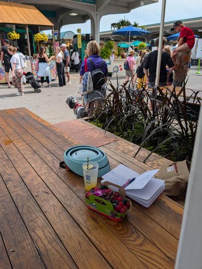 A photo of a picnic table with a basket of berries, a margarita, a journal and a bike helmet on it with people browsing a farmers market, a dj stand, and a woman and a dog in the background.