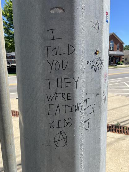 Graffiti written in black marker on the side of a metal lamp pole: “I told you they were eating kids” then an anarchist A in a circle.