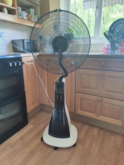 A tall misting fan stands in a kitchen. It's black and white with a control panel the space shuttle would be proud of. Maybe it's the hose but there's something a bit lifelike about it 🤨