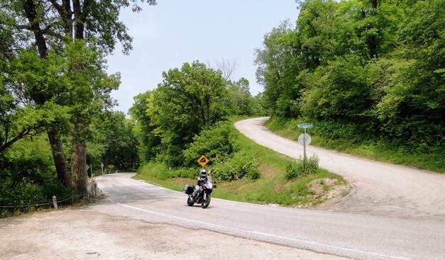 A motorcyclist in full riding gear waves toward the camera while riding down a winding, tree-lined rural road. The road curves through a hilly, densely wooded area with a steep downgrade warning sign visible nearby, and a green street sign reading "McClintock Rd" marks an intersecting gravel road at a stop sign.