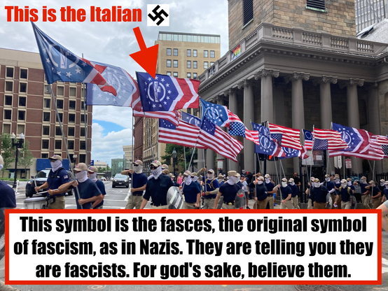 Photo of a group of masked marchers carrying inverted US, right-side-up 13-star US, but most importantly fascist flags with the fascia enclosed in a circle surrounded by stars on a blue field, with two right-pointing chevrons to its right in white on a red field. Along the top: "This is the Italian [swastika symbol]." Along the bottom, "This symbol is the fasces, the original symbol of fascism, as in Nazis. They are telling you they are fascists. For god's sake, believe them."