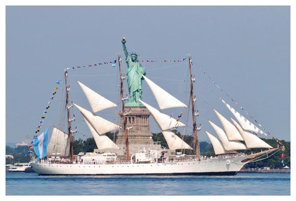 A long three-masted ship with triangular sails, and with small multicolored flags strung between the masts, passes in front of the Statue of Liberty. This is the Ara Libertad from Argentina, and it flies a large blue-and-white Argentinian flag at its rear.