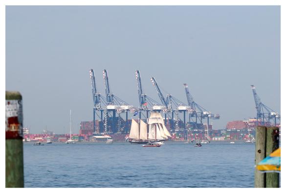 A three-masted tall ship with rectangular and triangular sails crosses the waters of New York Harbor, passing in front of a row of container cranes on the far shore. The image is framed by wooden posts at left and right, closer to the pier from which I’m taking the picture.