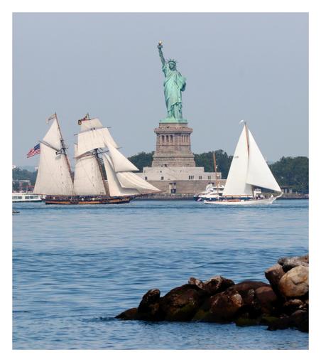 The Statue of Liberty is flanked by two sailing ships passing in front of it: to the left, a two-masted ship with rectangular and triangular sails (likely the American ship Pride of Baltimore II), and to the right, a smaller two-masted ship with triangular sails on the front mast and the sails on the back mast furled, which I can’t quite identify. In the foreground, a low pile of rocks pushes into the waters of the bay.