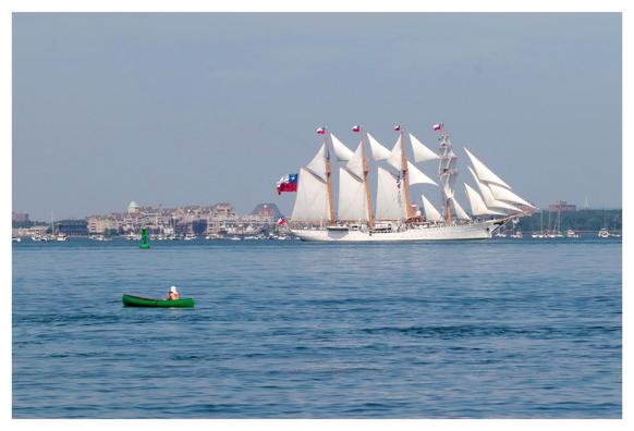 The four-masted ship Esmeralda from Chile, with multiple rectangular and triangular sails, moves through the waters of New York Harbor under a cloudless sky. Floating some distance in front of it is a tiny green canoe, in which a shirtless person sits with their head wrapped in white cloth, observing the scene.