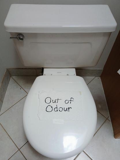 A small toilet with the over down and a piece of paper towel taped on it which reads, "Out of Odour".