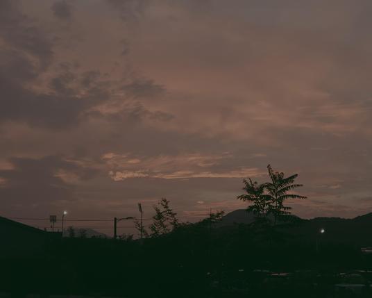 A photograph of a cloudy sky featuring shades of muted purple, gray, and faint pink. The expanse of the sky is filled with various cloud formations. formations. Below the horizon line, dark silhouettes of mountains and trees are visible. A utility pole with wires and small points of light appear along the darkened skyline. The bottom foreground of the image consists of deep shadows.