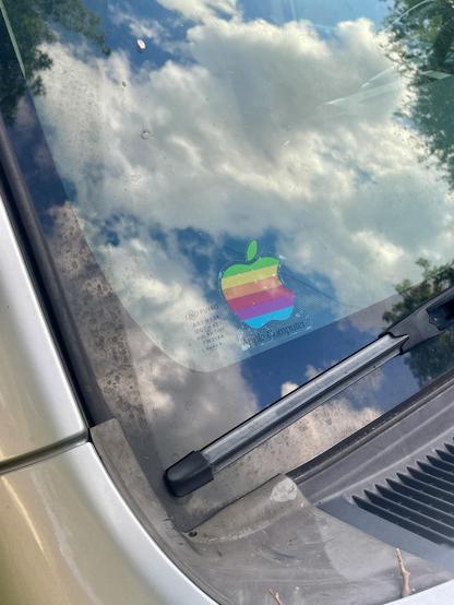 A 1990’s era Apple rainbow logo sticker with a transparent background on the corner of my car’s windshield. The windshield is reflecting a partly cloudy sky with some dramatic clouds.