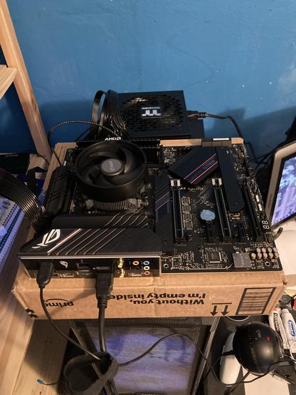A computer motherboard, with some necessary components installed, on a cardboard box on top of an active PC on top of a filing cabinet. A power supply rests behind it. This is, in fact, a minimum viable PC. You can actually build a PC on a cardboard box and it will function just fine. It's not recommended to put a full weight GPU onto something like this, however, as it can damage the expansion card slot.